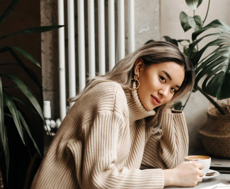 blonde asian girl in beige oversized sweater sits in cafe with cup of coffee and carrot juice.